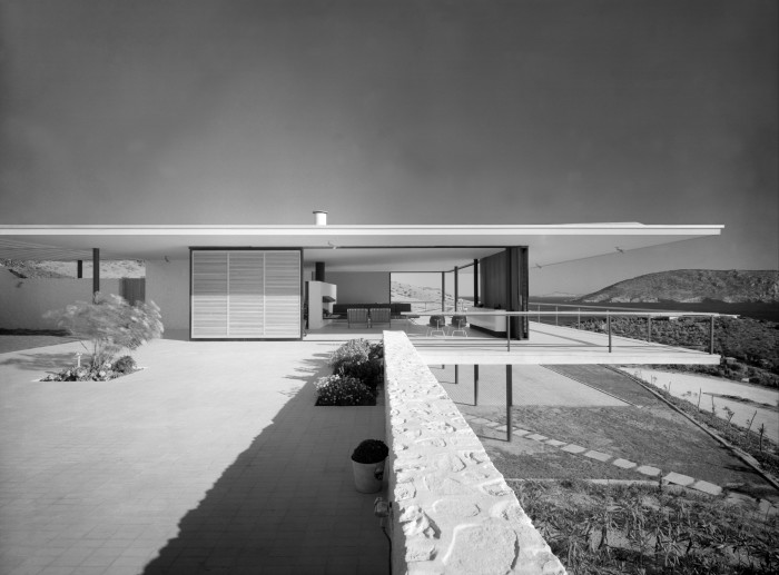 Black and white photograph of a very modern-looking one-story building with a flat overhanging roof resting on several thin pillars 